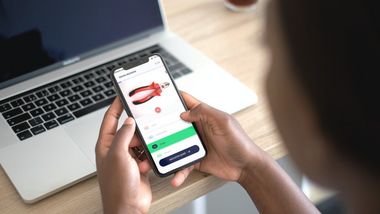 Eine Person sitzt vor dem Laptop mit einem Handy in den Händen. Auf dem Handy ist die Lunes App geöffnet und die Abbildung einer roten Zange. Darunter in einer Liste das Wort Zange ausgewählt.