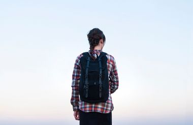 Frau mit geflochtenem Haar, Karohemd und schwarzem Rucksack steht mit dem Rücken zur Kamera vor einem hellen Himmel.